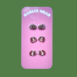 Könnte beinhalten: Eine rosa Karte mit dem Text "GARLIC HEAD" in weißen Buchstaben. Die Karte zeigt drei Paare von Meerschweinchen-Ohrringen. Das erste Paar ist braun und weiß, das zweite Paar ist braun und weiß und das dritte Paar ist grau und weiß.