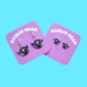 Puede incluir: Un par de pendientes de aro de plata con un diseño de cara de lémur en una tarjeta rosa con el texto "GARLIC HEAD" en letras blancas.