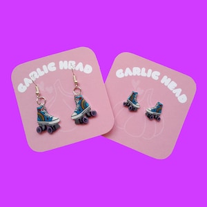 Op de afbeelding: Een paar oorbellen met een blauw rolstoeltje ontwerp met regenboog accenten. De oorbellen zijn op een roze achtergrond met de tekst "GARLIC HEAD" gedrukt op elke kaart.