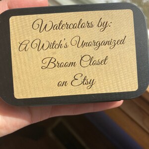 May include: A black box with a gold label that reads "Watercolors by: A Witch's Unorganized Broom Closet on Etsy".