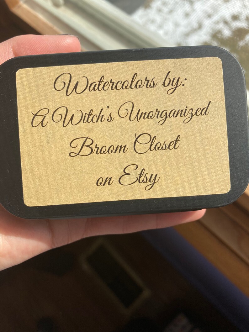 May include: A black box with a gold label that reads "Watercolors by: A Witch's Unorganized Broom Closet on Etsy".