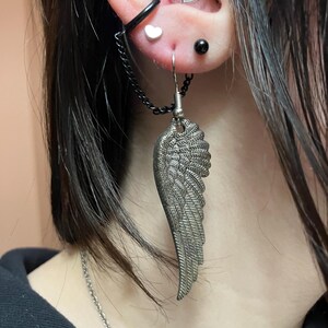 May include: A silver wing-shaped earring with a chain attached to a black hoop earring. The earring is worn in the upper lobe of the ear.