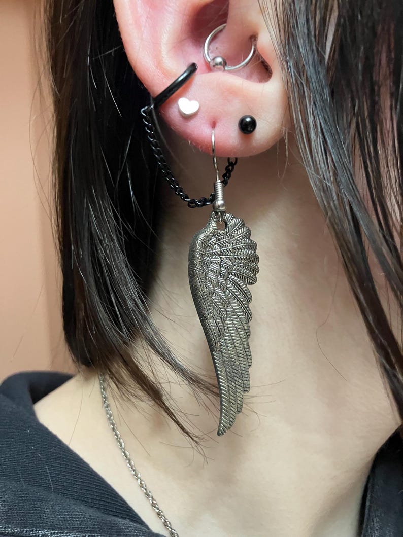 May include: A silver wing-shaped earring with a chain attached to a black hoop earring. The earring is worn in the upper lobe of the ear.