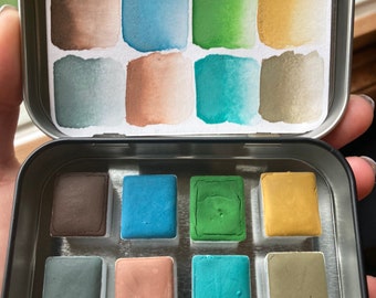 May include: A small, open metal tin containing watercolor paints. The tin holds eight square paint pans in various colors, including brown, blue, green, and gold. The lid of the tin has six rectangular watercolor swatches in similar hues.