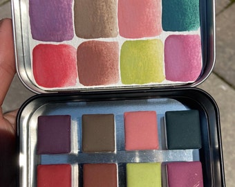 Könnte beinhalten: Eine Metalldose mit einem Satz von 12 Aquarellfarben. Die Farben sind in zwei Reihen mit jeweils sechs Farben angeordnet, wobei jede Farbe in einer quadratischen Schale liegt. Die Farben umfassen Lila, Braun, Rosa, Grün, Rot und Gelb.