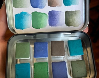 Könnte beinhalten: Eine kleine, offene Metallbox mit acht Aquarellfarbenquadraten in Blau-, Grün- und Brauntönen. Der Deckel der Dose zeigt Farbmuster in den gleichen Farben. Die Farben sind in zwei Reihen zu je vier Stück angeordnet.