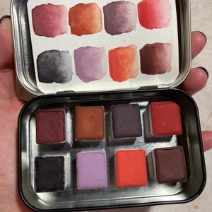 May include: A metal tin with a set of 12 watercolor paint pans. The pans are arranged in a 3x4 grid and are filled with various shades of red, orange, purple, and brown.