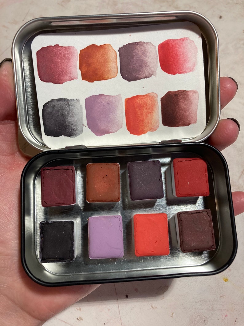 May include: A metal tin with a set of 12 watercolor paint pans. The pans are arranged in a 3x4 grid and are filled with various shades of red, orange, purple, and brown.