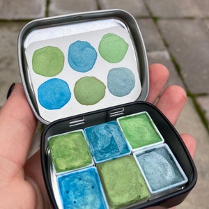 May include: A metal tin with a set of eight watercolor paints. The paints are arranged in a grid pattern and are in shades of green and blue.