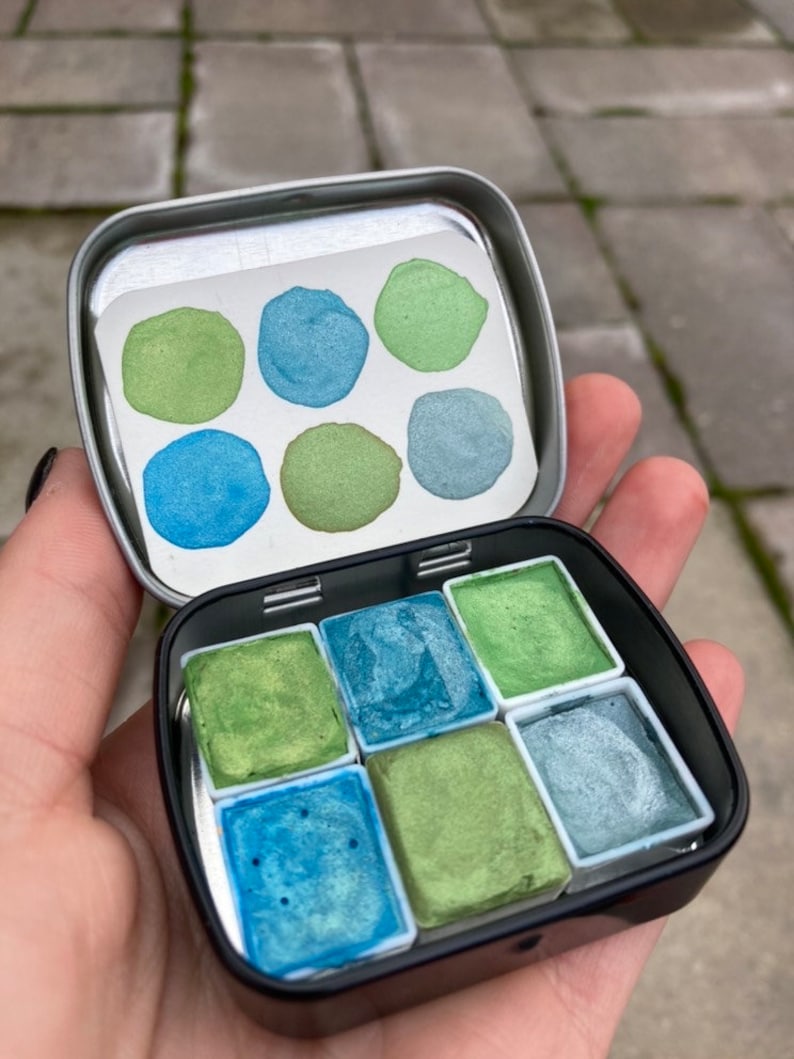 May include: A metal tin with a set of eight watercolor paints. The paints are arranged in a grid pattern and are in shades of green and blue.