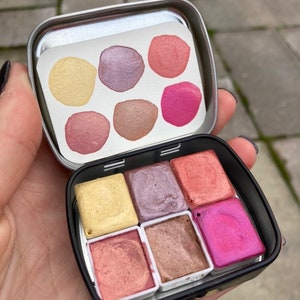 May include: A metal tin with six watercolor paint pans. The pans are arranged in two rows of three. The colors are yellow, purple, pink, peach, brown, and magenta.