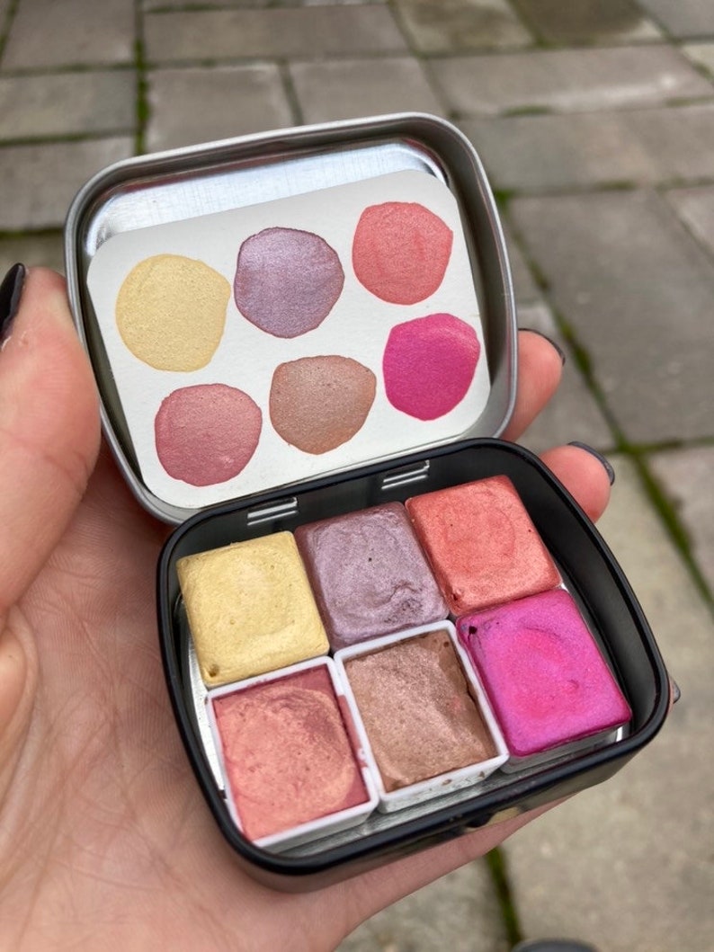 May include: A metal tin with six watercolor paint pans. The pans are arranged in two rows of three. The colors are yellow, purple, pink, peach, brown, and magenta.