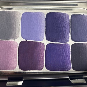 K&ouml;nnte beinhalten: Eine Metallpalette mit acht Aquarellfarben-Schalen. Die Schalen sind mit Farbt&ouml;nen in Lila, Blau und Grau gef&uuml;llt.