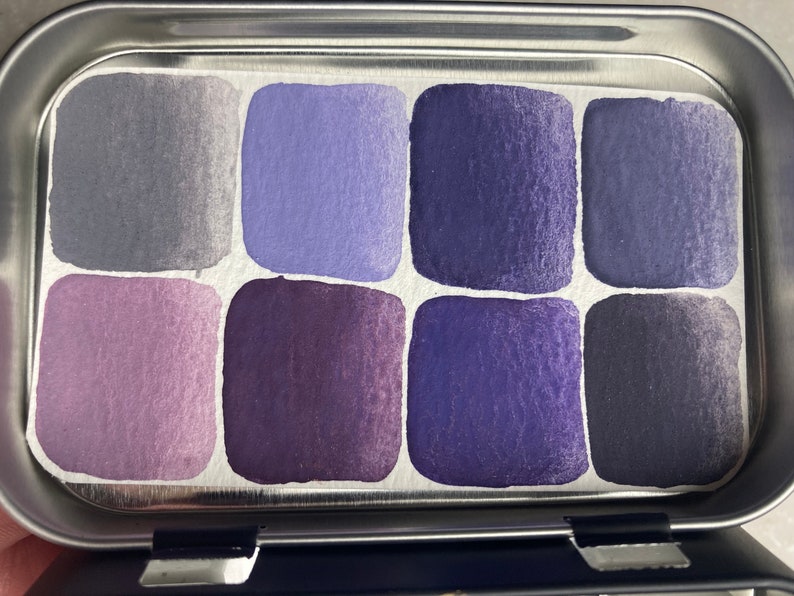 K&ouml;nnte beinhalten: Eine Metallpalette mit acht Aquarellfarben-Schalen. Die Schalen sind mit Farbt&ouml;nen in Lila, Blau und Grau gef&uuml;llt.