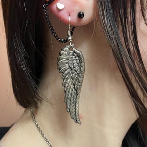May include: A silver angel wing earring with a chain attached to a small black hoop. The earring is worn in the earlobe and the chain goes up to the helix piercing.