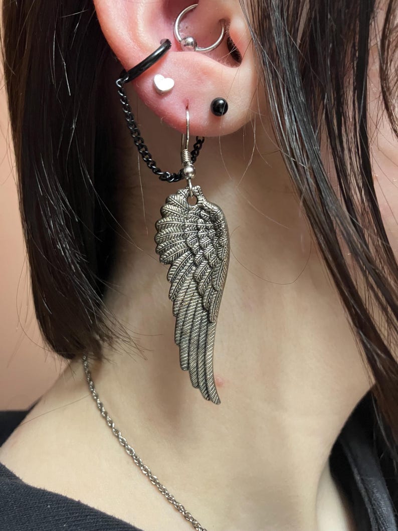 May include: A silver angel wing earring with a chain attached to a small black hoop. The earring is worn in the earlobe and the chain goes up to the helix piercing.