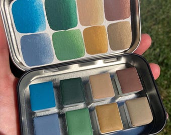May include: A metal tin with a removable lid containing 12 watercolor paint pans. The pans are arranged in two rows of six. The colors are blue, green, brown, and beige.