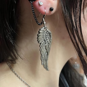 May include: A silver angel wing earring with a chain attached to a black hoop earring. The earring is worn in the ear of a person with black hair.