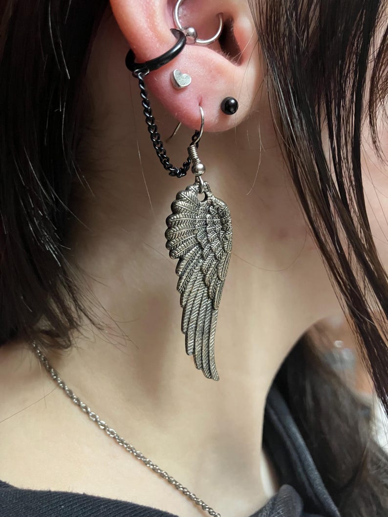 May include: A silver angel wing earring with a chain attached to a black hoop earring. The earring is worn in the ear of a person with black hair.