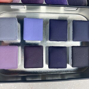 K&ouml;nnte beinhalten: Eine Metalldose mit neun Aquarellfarben. Die Farben sind in drei Reihen zu je drei angeordnet. Die Farben sind in verschiedenen Schattierungen von Lila und Blau.