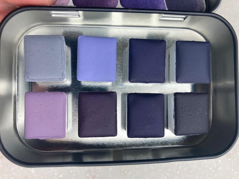 K&ouml;nnte beinhalten: Eine Metalldose mit neun Aquarellfarben. Die Farben sind in drei Reihen zu je drei angeordnet. Die Farben sind in verschiedenen Schattierungen von Lila und Blau.