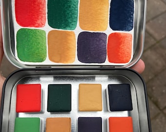 May include: A metal tin with a set of 9 watercolor paint pans. The pans are arranged in a 3x3 grid and are filled with red, green, yellow, blue, orange, and purple paint.