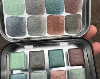 May include: A metal tin with a set of 12 watercolor paints. The paints are arranged in a grid of four rows and three columns. The colors are a mix of greens, blues, and browns.