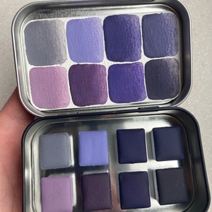 K&ouml;nnte beinhalten: Eine Metalldose mit einem Satz von 12 Aquarellfarben. Die Farben sind in zwei Reihen mit jeweils sechs Farben angeordnet. Die Farben sind in verschiedenen Schattierungen von Lila und Blau.