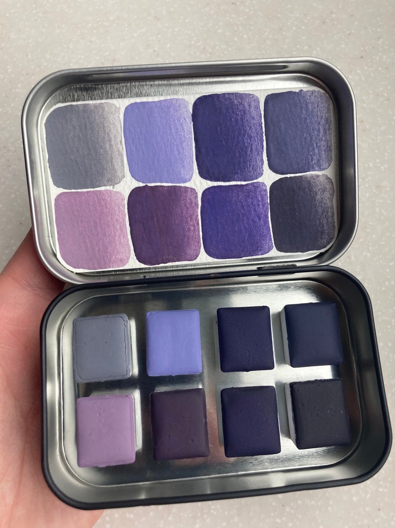 K&ouml;nnte beinhalten: Eine Metalldose mit einem Satz von 12 Aquarellfarben. Die Farben sind in zwei Reihen mit jeweils sechs Farben angeordnet. Die Farben sind in verschiedenen Schattierungen von Lila und Blau.