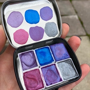 May include: A metal tin with a set of eight watercolor paints. The paints are in a variety of shades of purple, blue, and silver. The tin is open and the paints are visible.