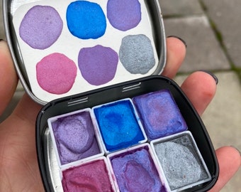 May include: A metal tin with a set of eight watercolor paints. The paints are in a variety of shades of purple, blue, and silver. The tin is open and the paints are visible.