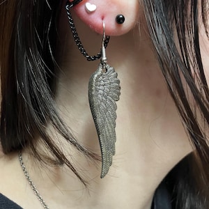 May include: A silver wing-shaped earring with a chain attached to a hoop earring. The earring is hanging from a person's ear, which is pierced multiple times.