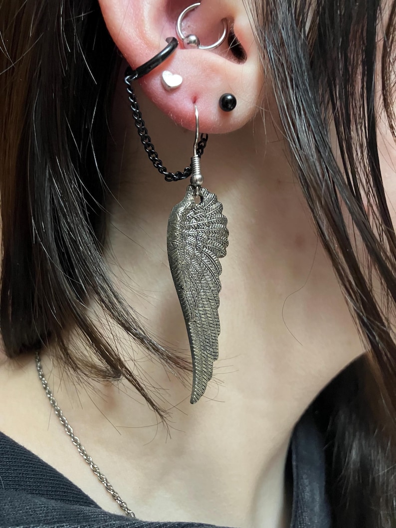 May include: A silver wing-shaped earring with a chain attached to a hoop earring. The earring is hanging from a person's ear, which is pierced multiple times.