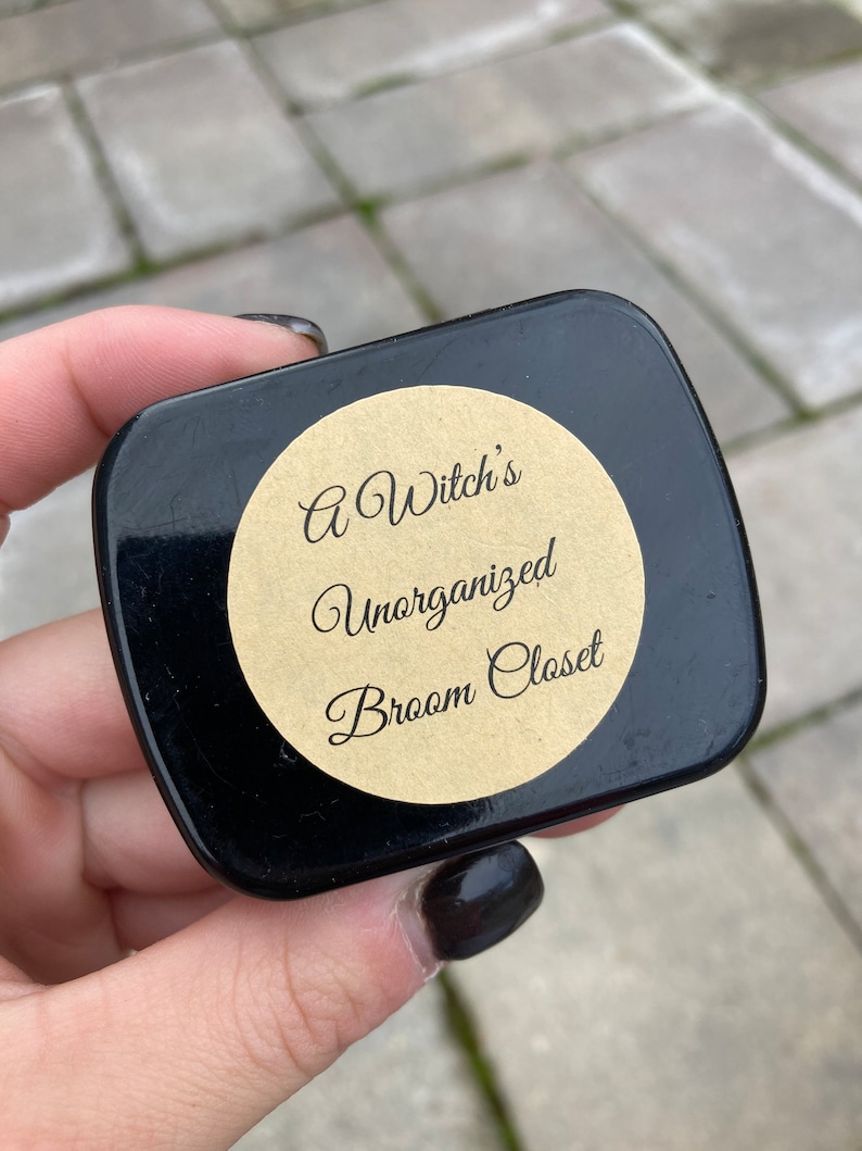 May include: A black round tin with a brown paper label that reads "A Witch's Unorganized Broom Closet".