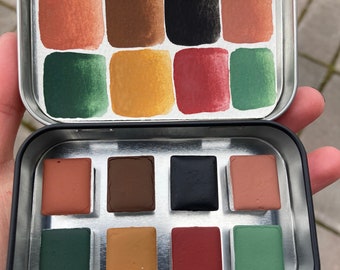 Könnte beinhalten: Eine Metalldose mit einem Satz von 12 Aquarellfarben. Die Farben sind in einem Raster von vier Reihen und drei Spalten angeordnet. Die Farben umfassen Braun-, Grün-, Rot- und Orangetöne.