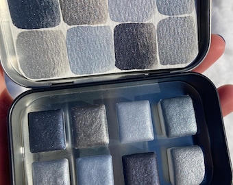 May include: A metal tin with a clear lid containing a set of nine watercolor paints in shades of gray and silver. The paints are arranged in a 3x3 grid.
