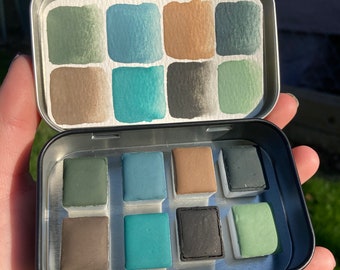 Könnte beinhalten: Eine Metalldose mit einem Satz von 12 Aquarellfarben in Grüntönen, Blau, Braun und Schwarz. Die Farben sind in einem Raster angeordnet und bereit zum Malen.