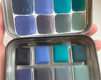 Könnte beinhalten: Eine Metalldose mit einem Satz von 12 Aquarellfarben in Blautönen, Grüntönen und Grautönen. Die Farben sind in zwei Reihen zu sechs angeordnet, wobei die obere Reihe die Farben in einer satteren Form zeigt und die untere Reihe die Farben in einer gedämpfteren Form zeigt.