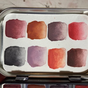 May include: A metal tin with a watercolor paint palette inside. The palette has eight squares of paint in shades of red, orange, brown, purple, and gray.