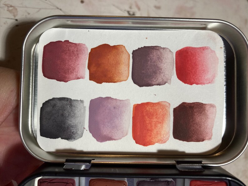 May include: A metal tin with a watercolor paint palette inside. The palette has eight squares of paint in shades of red, orange, brown, purple, and gray.