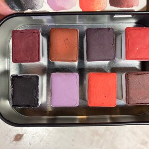 May include: A metal tin with eight watercolor paint pans. The pans are arranged in two rows of four. The colors are red, orange, purple, black, brown, and a lighter shade of brown.