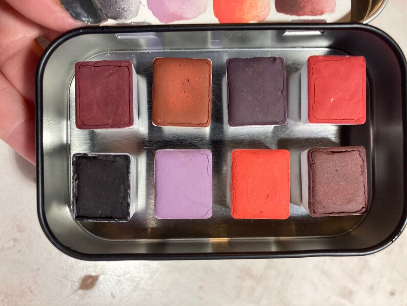 May include: A metal tin with eight watercolor paint pans. The pans are arranged in two rows of four. The colors are red, orange, purple, black, brown, and a lighter shade of brown.