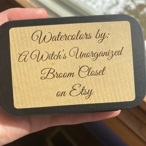 K&ouml;nnte beinhalten: Eine schwarze Dose mit einem braunen Papieretikett, auf dem "Watercolors by: A Witch's Unorganized Broom Closet on Etsy" steht.