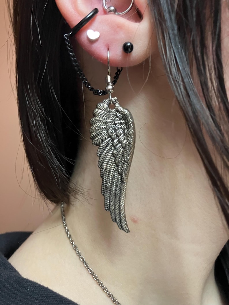 May include: A silver wing-shaped earring with a chain attached to a black hoop earring. The earring is worn in the earlobe and the hoop is worn in the upper ear.