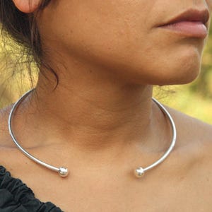 Puede incluir: Un collar gargantilla plateado con un diseño minimalista. El collar es una barra curva rígida que se apoya alrededor del cuello, con una pequeña bola en cada extremo. La joya se lleva en el cuello de una persona.