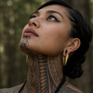 May include: A pair of gold-toned ear weights featuring a sleek, elongated triangular shape with a central vertical cutout. These metallic ornaments are designed to be worn through stretched earlobes, showcasing a polished finish and a sharp, pointed base.