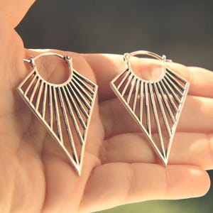Könnte beinhalten: Ein Paar silberfarbene Ohrringe. Jeder Ohrring hat ein geometrisches, offenes Design mit einer rautenförmigen Form und einem Sonnenstrahlenmuster. Die Ohrringe werden in einer Hand vor einem verschwommenen Hintergrund gehalten.