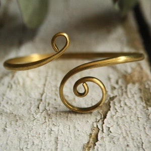 Könnte beinhalten: Ein goldfarbenes Metallarmband mit einem zarten, wirbelnden Design. Das Armband ist offen und verstellbar.