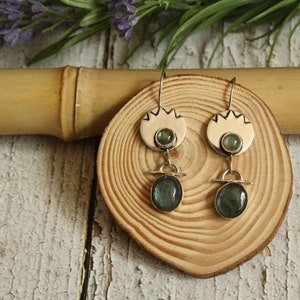 May include: A pair of silver earrings with a round, white, textured top and a dangling oval-shaped green gemstone. The earrings are displayed on a wooden surface.