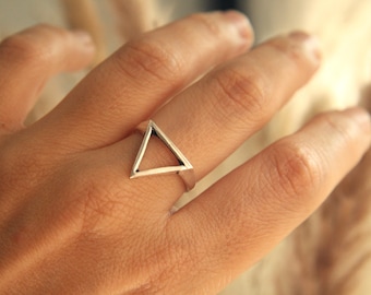 May include: A silver ring featuring a hollow triangle design. The geometric ring is worn on a finger, showcasing its minimalist and modern aesthetic. The ring's clean lines and open shape create a striking visual contrast.